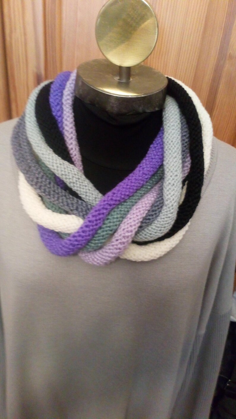 Pu&ograve; includere: Una sciarpa infinita di maglia multicolore con sfumature di viola, bianco, grigio e nero.