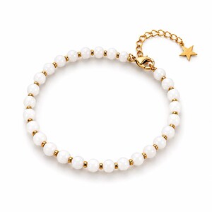Peut inclure: Un bracelet délicat composé de perles blanches rondes, séparées par de petits éléments dorés. Il est orné d'une chaîne et d'un fermoir dorés, ainsi que d'un petit charme en forme d'étoile. Le bijou est présenté sur fond blanc.