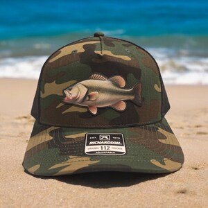 May include: Camouflage trucker hat with a large fish patch. The hat has a black mesh back and a curved brim. The fish is a light brown color with red accents. The hat has a Richardson 112 Trucker patch.
