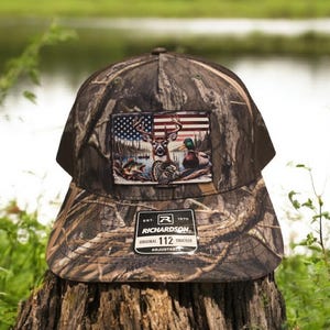 Peut inclure: Un chapeau de camionneur camouflage avec un patch repr&eacute;sentant un cerf, des canards et un drapeau am&eacute;ricain. Le chapeau est r&eacute;glable et porte le texte "Richardson 112" sur le devant.