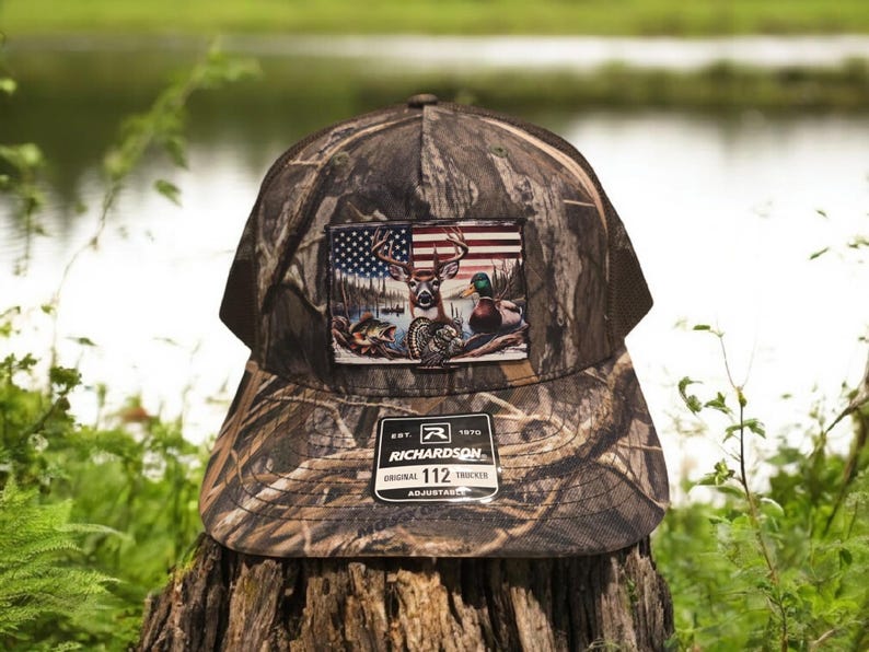 Peut inclure: Un chapeau de camionneur camouflage avec un patch repr&eacute;sentant un cerf, des canards et un drapeau am&eacute;ricain. Le chapeau est r&eacute;glable et porte le texte "Richardson 112" sur le devant.