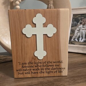 May include: Wooden block with a light-colored cross cutout. The block has a light brown wood grain and includes the text: "I am the light of the world. Anyone who follows me will never walk in the darkness but will have the light of life."