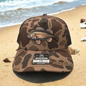 May include: A brown camouflage trucker hat with a fish graphic. The hat features a mesh back and a curved brim. The front of the hat displays a detailed illustration of a fish. The hat has a Richardson 112 Trucker tag.
