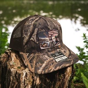 Peut inclure: Un chapeau de camionneur camouflage brun et vert avec un patch repr&eacute;sentant un cerf, un canard et un drapeau am&eacute;ricain. Le chapeau est fabriqu&eacute; par Richardson 112 et est r&eacute;glable.
