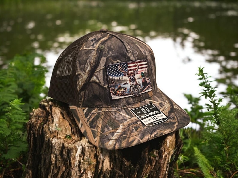 Peut inclure: Un chapeau de camionneur camouflage brun et vert avec un patch repr&eacute;sentant un cerf, un canard et un drapeau am&eacute;ricain. Le chapeau est fabriqu&eacute; par Richardson 112 et est r&eacute;glable.