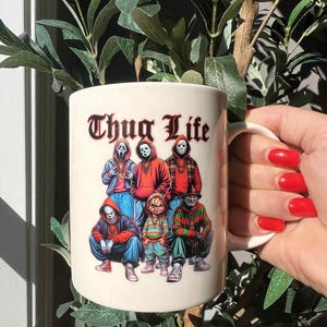 May include: A white ceramic mug featuring the text "Thug Life" in a gothic font above a graphic of six horror movie characters. The characters are wearing hoodies, and one is wearing a mask.