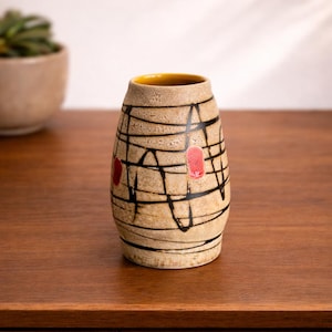Könnte beinhalten: Eine beige Keramikvase mit gelbem Innenraum und strukturierter Oberfläche. Die Vase ist mit schwarzen Linien und zwei roten Akzenten verziert. Die Vase steht auf einer Holzoberfläche.