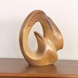 Può includere: Un vaso in ceramica con una forma scultorea astratta a ciclo aperto. La superficie presenta una sfumatura di caldi toni marrone chiaro e beige, accentuata da uno smalto testurizzato con macchie marrone scuro lungo i bordi e l'elemento centrale.
