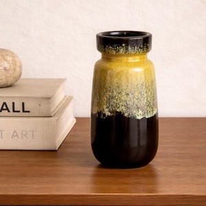 Könnte beinhalten: Eine weiße Keramik-Tasse mit schwarzem Henkel und dem Text "But First, Coffee" in fetter, schwarzer Schrift. Die Tasse steht vor einem verschwommenen Hintergrund, der ihr schlichtes Design hervorhebt.