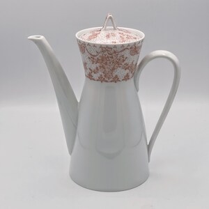 Könnte beinhalten: Weiße Kaffeekanne aus Keramik mit braunem Blumendesign am oberen Rand. Die Kanne hat einen Deckel und einen Henkel.