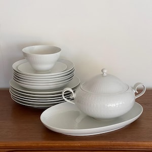 Könnte beinhalten: Ein weißes Porzellan-Geschirrset, bestehend aus Tellern, Schalen und einer Servierschüssel mit Deckel. Das Set hat ein strukturiertes Muster und ist auf einer Holzoberfläche angeordnet. Die Terrine steht auf einer ovalen Platte.