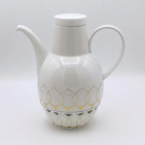 Op de afbeelding: Witte keramische koffiepot met een deksel, gebogen tuit en handvat. De pot heeft een gouden geometrisch patroon rond de basis. Het ontwerp is elegant en geschikt voor het serveren van koffie of thee.
