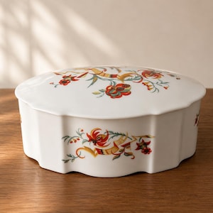 Può includere: Un portagioie in ceramica bianca con coperchio, con un motivo floreale rosso, giallo e verde. La scatola ha un bordo smerlato e poggia su una superficie di legno. Il motivo floreale è ripetuto sul coperchio e sui lati.