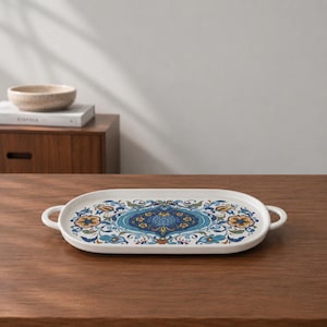 Könnte beinhalten: Ein ovales Serviertablett aus weißer Keramik mit zwei seitlichen Griffen, das in der Mitte ein blaues, gelbes und grünes Blumenmuster mit komplexen symmetrischen Designs und geschwungenen Blattmotiven auf der flachen Oberfläche zeigt.