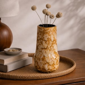Op de afbeelding: Een decoratieve keramische vaas met een getextureerd, gevlekt patroon in tinten geel en crème. De vaas bevat gedroogde bloemstelen. Hij staat op een geweven dienblad op een houten oppervlak, naast boeken en een klein schaaltje.