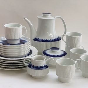 Op de afbeelding: Een wit porseleinen theeservies met blauwe en witte geometrische ontwerpen. De set bevat een theepot, suikerpot, roomkan en zes kopjes en schoteltjes.