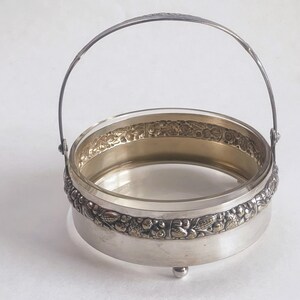 Op de afbeelding: Zilveren serveermand met een heldere glazen kom. De mand heeft een decoratief bloemen- en bladerpatroon rond de basis en een handvat. De mand staat op drie kleine voetjes.