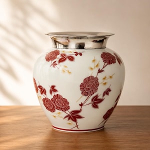 Puede incluir: Un jarrón de cerámica blanca con borde y base plateados. El jarrón está decorado con un estampado floral en tonos rojos y amarillos. El jarrón está sobre una superficie de madera.