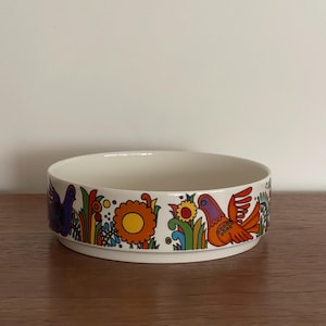 Könnte beinhalten: Eine weiße Keramikschale mit einem farbenfrohen Blumen- und Vogelmuster. Der Rand zeigt orange, gelbe, blaue und rote Vögel und Blumen. Die Schale ist rund und steht auf einer Holzoberfläche.