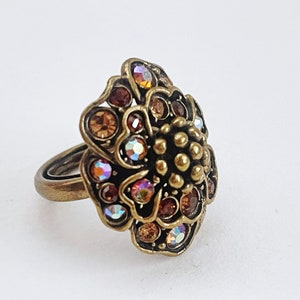 Könnte beinhalten: Ein goldfarbener Ring mit einem großen blütenförmigen Design. Die Blume ist mit schillernden und bernsteinfarbenen Strasssteinen verziert.