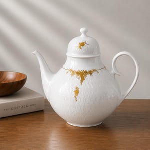 Könnte beinhalten: Eine weiße Porzellanteekanne mit einem dezenten, geprägten Ovalmuster auf der Oberfläche, verziert mit zarten goldenen Blumenmotiven auf dem Körper und dem Deckel. Die Teekanne verfügt über einen geschwungenen Henkel und einen Deckel.