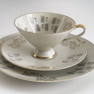 May include: A white porcelain teacup and saucer set with a gold trim and a floral pattern. The cup has a wide, shallow bowl and a handle. The saucer has a matching pattern and a slightly raised edge.
