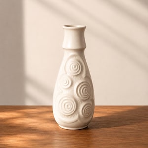 Könnte beinhalten: Eine cremefarbene Keramikvase mit bauchigem Körper und zylindrischem Hals. Die Vase hat ein sich wiederholendes Muster aus geprägten konzentrischen Kreisen. Die Vase steht auf einer Holzoberfläche.