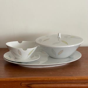 Könnte beinhalten: Ein weißes Keramik-Servier-Set mit einer großen Schüssel mit Deckel, einer kleineren Schüssel und einer rechteckigen Platte. Das Set ist mit einem Feder-Muster in gedämpften Farben verziert. Die Schüsseln und die Platte stehen auf einer Holzoberfläche.