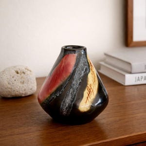 Könnte beinhalten: Eine schwarze Keramikvase mit einer einzigartigen Form und einem farbenfrohen abstrakten Design. Die Vase hat rote und gelbe Pinselstriche auf dunklem Hintergrund. Ein dekoratives Stück für die Inneneinrichtung.