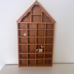 May include: A wooden display shelf, house-shaped at the top, with numerous small square compartments. The shelf is made of light brown wood and contains a small ceramic bunny figurine and a coin.