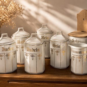 Può includere: Una collezione di barattoli da cucina in ceramica bianca e una brocca, con scritte dorate e dettagli decorativi. I barattoli sono etichettati con termini alimentari tedeschi. È inclusa anche una saliera in legno.