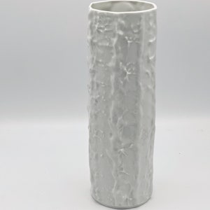 Könnte beinhalten: Weiße Keramikvase mit einem strukturierten, baumrindenartigen Design. Die Vase ist hoch und zylindrisch.