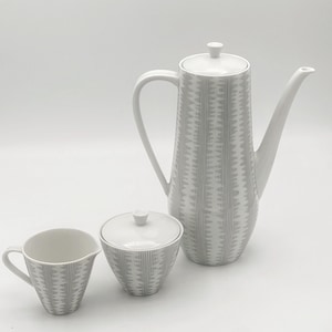 Op de afbeelding: Een witte keramische koffieset met een grijs geometrisch patroon. De set bevat een hoge koffiepot, een melkkan en een suikerpot met een deksel. De koffiepot heeft een lange tuit en een gebogen handvat. De melkkan heeft een klein handvat en de suikerpot is rond.