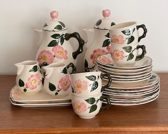 Op de afbeelding: Een keramieken theeservies bestaande uit twee theepotten met deksels, twee kannetjes, vier theekopjes en een stapel schoteltjes. Elk stuk is crèmekleurig met handgeschilderde roze bloemen, groene bladeren en donkergroene oren.