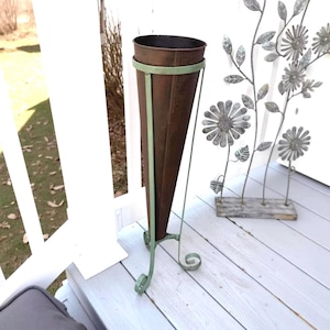 May include: A tall, cone-shaped brown metal planter held by a light green metal stand with three curved legs. The planter has a wide circular opening at the top and a tapered base, supported by a ring frame that connects to the legs.