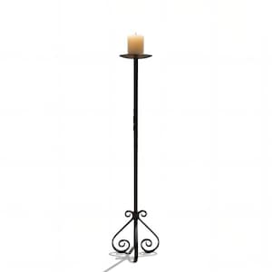 May include: A tall, slender black metal floor candle holder featuring a circular top plate and a decorative base with curved, scroll-like metalwork. A single, cylindrical off-white pillar candle sits centered on the top plate.