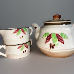 May include: A white ceramic tea set with a teapot and two stacked cups. The teapot has a brown lid and spout. The cups and teapot are decorated with red, green, and brown leaf designs. The set is on a light gray surface.