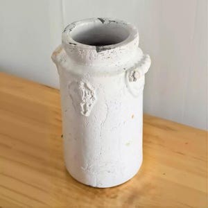 May include: A cylindrical white vase with a distressed, crackled finish and a textured surface. The vase features a wide, thick rim and two small, molded side handles near the top. The exterior shows subtle signs of wear and uneven paint application.