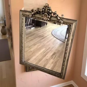 May include: A rectangular wall mirror featuring an ornate, metallic gold-toned frame with intricate scrollwork and a shell-like motif at the top center. The frame has a beaded inner edge and a smooth, reflective glass surface that shows a room reflection.