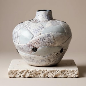 May include: A handcrafted ceramic vase with a patchwork design. The vase features a rounded body and a narrow neck, with a surface adorned with various textured patches in shades of white, gray, and blue. Two black circular accents are visible on the vase.