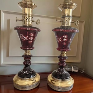 May include: Pair of antique ruby red glass lamps with brass accents. The lamps feature a cut glass design, ornate brass fittings, and a decorative base. The lamps are displayed on a wooden surface.