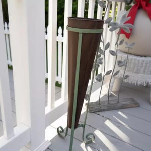 May include: A tall, brown metal planter with a conical shape, accented with light green trim and a decorative metal stand. The planter is set on a wooden deck next to a white picket fence.