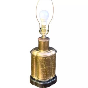 May include: A brass table lamp with a cylindrical body featuring etched floral and bird designs. The lamp has a dark base, a brass neck, a metal harp, and a single white light bulb. The metal surface shows a polished finish with detailed engravings.