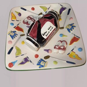 May include: A square, white ceramic golf-themed plate with a green border. The plate features a golf bag, golf shoes, golf balls, flags, and golf clubs. The golf bag is black and burgundy. The plate has a whimsical design.