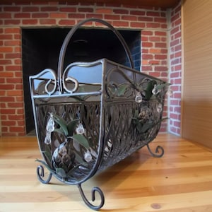 May include: A decorative metal magazine rack with a handle, featuring a woven basket design. The rack is adorned with leaf and crystal accents, and rests on scrolled feet. The color is a dark bronze.