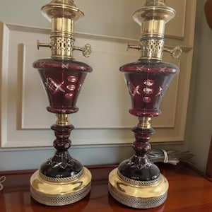 May include: Two antique oil lamps with ruby red glass bodies and brass accents. The lamps have a decorative etched design on the glass and sit on a gold-colored base. The lamps are displayed on a wooden surface.