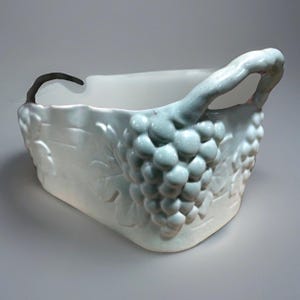 May include: A rectangular, off-white ceramic basket with a handle and grape cluster design. The handle is a curved stem shape, and the grapes are a light blue-green color. The basket has a textured surface and is likely for decorative use.