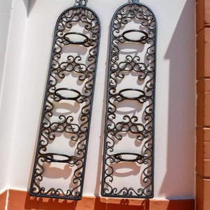 May include: Two black wrought iron wall-mounted wine racks. Each rack has an arched top and holds four bottles. The design features decorative scrollwork and oval bottle holders. The racks are mounted on a white wall with a red-tiled base.