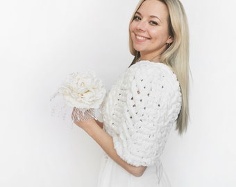 May include: A white chunky knit bolero with a textured cable pattern and short sleeves, paired with a decorative white fabric flower accessory featuring delicate dangling beads and a fine mesh netting detail on a white background.