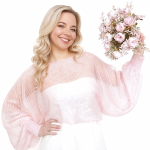 May include: A woman wearing a light pink, open-weave sweater with long, voluminous sleeves. She is holding a bouquet of pink flowers. The sweater has a cropped design and is paired with white clothing. The woman is smiling.